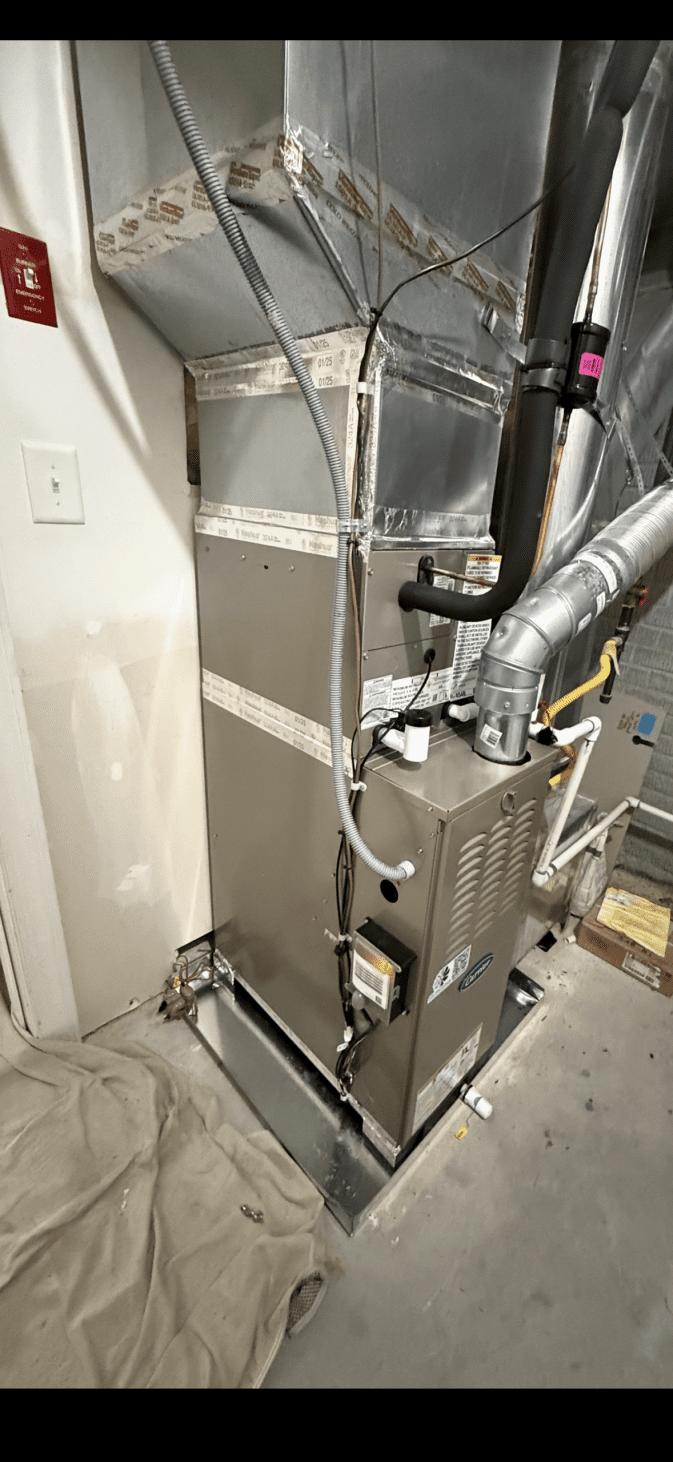 A metal furnace unit connected to ductwork stands on a basement floor near a wall, with various pipes, wires, and a beige cloth on the ground beside it—offering a closer look at the equipment featured in our About Us section.