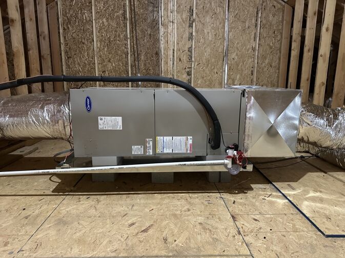 A large HVAC air handler unit with ductwork, detailed in our About Us section, is installed in an unfinished attic with wooden framing and plywood flooring.