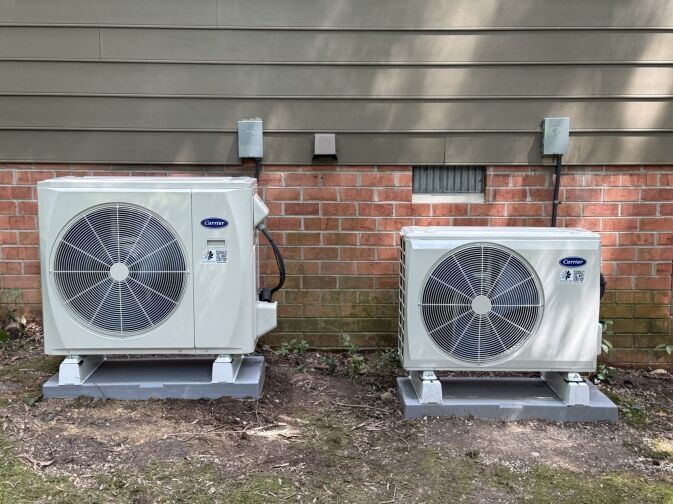 About Us: Two outdoor Carrier HVAC units installed on concrete pads beside a brick and siding house wall, each connected to electrical boxes.