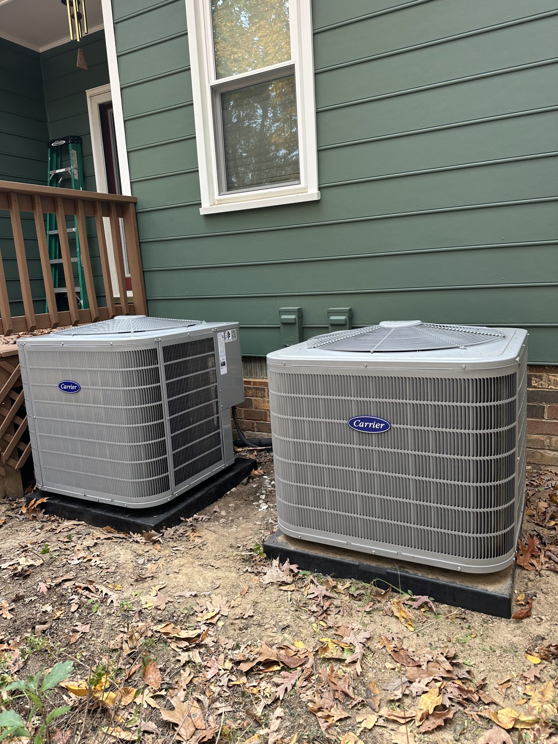 Two outdoor Carrier air conditioning units are installed on concrete pads next to a green house with a wooden deck and window above. Learn more About Us and our commitment to quality installation for your home comfort needs.