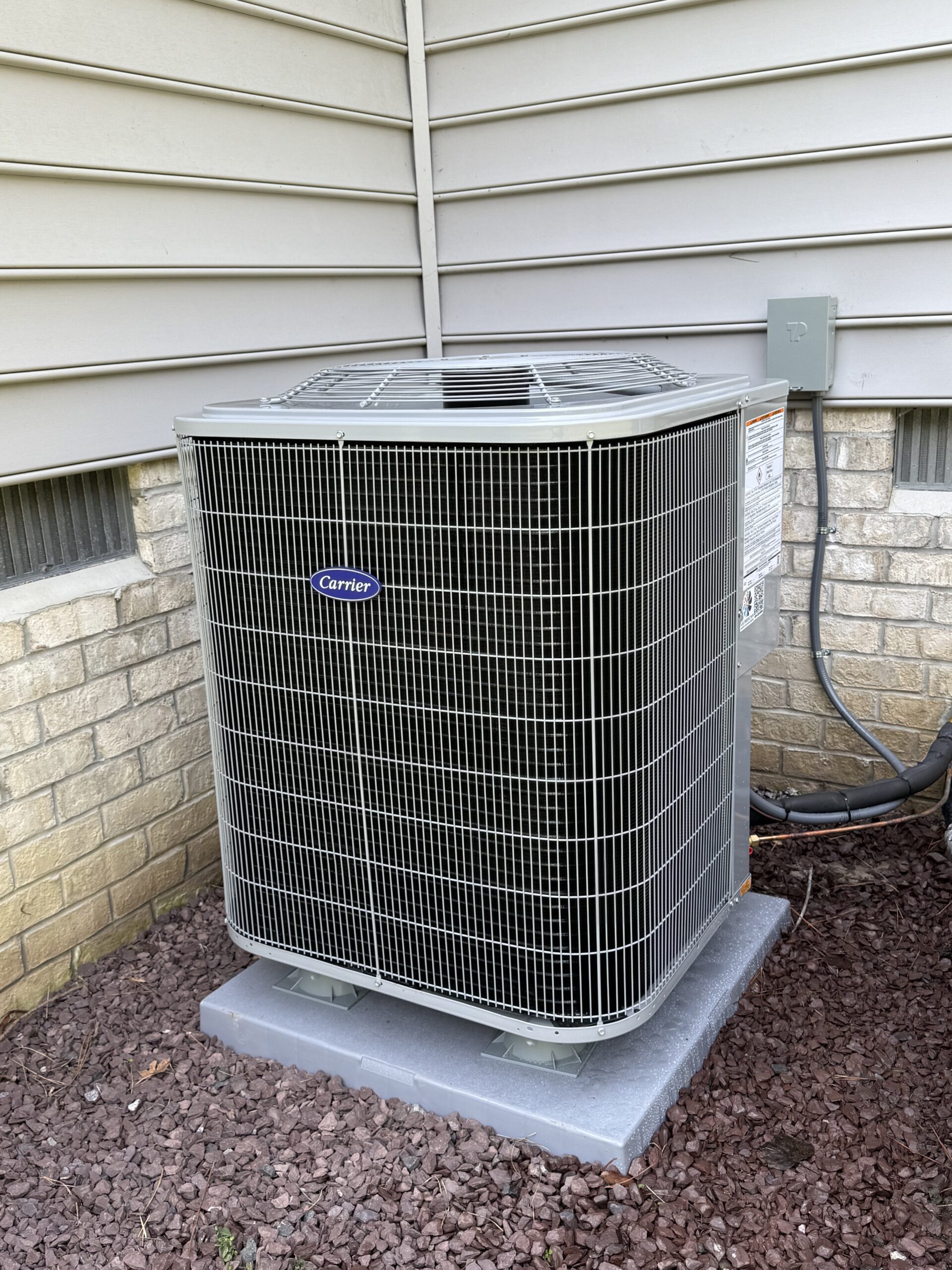 About Us: Outdoor Carrier air conditioning unit installed on a concrete pad next to a house with brick and siding exterior.