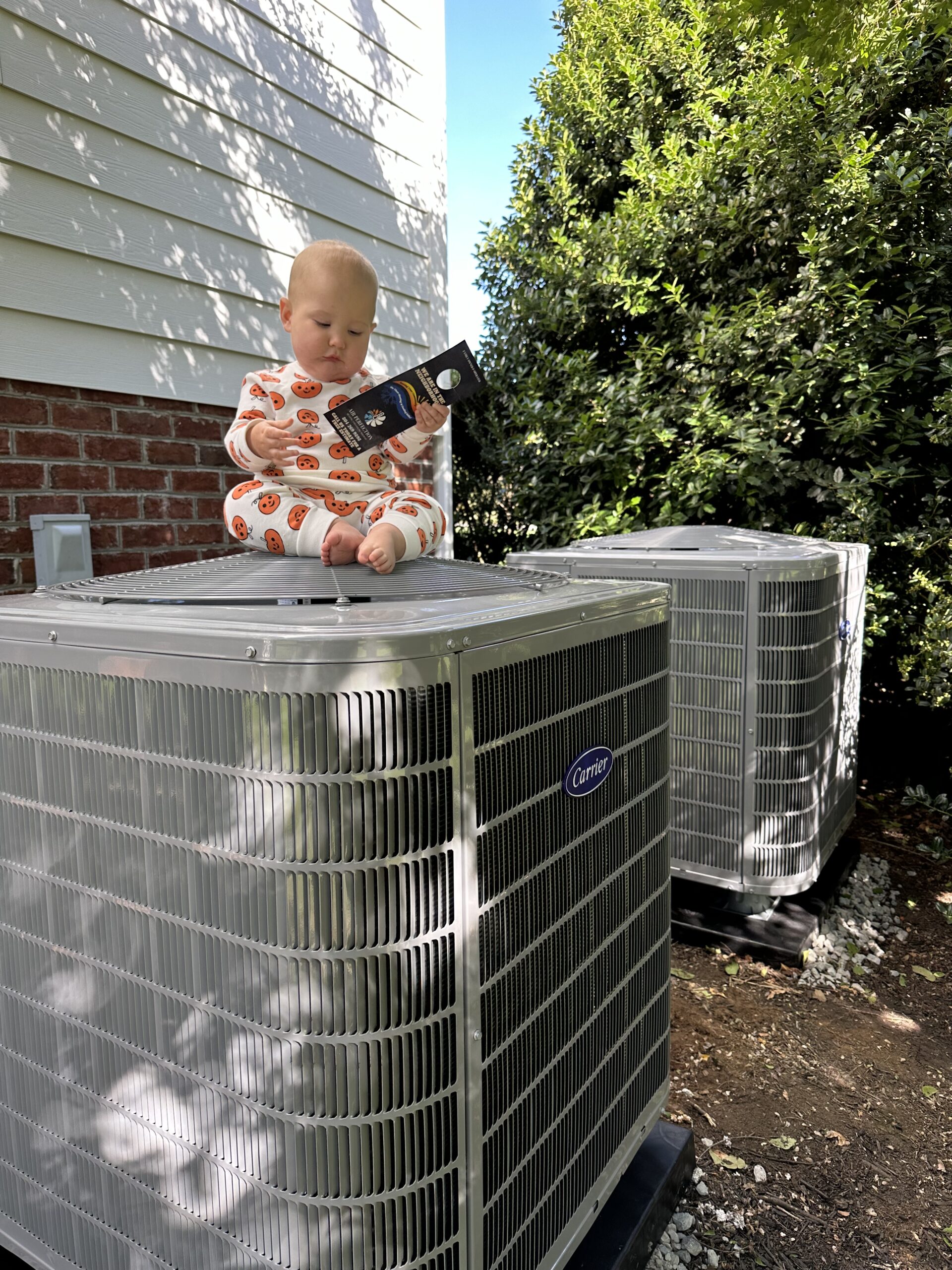 A baby in patterned pajamas sits atop an outdoor air conditioning unit, holding and looking at a smartphone. Nearby, another unit and greenery are visible, adding context to this candid About Us moment.