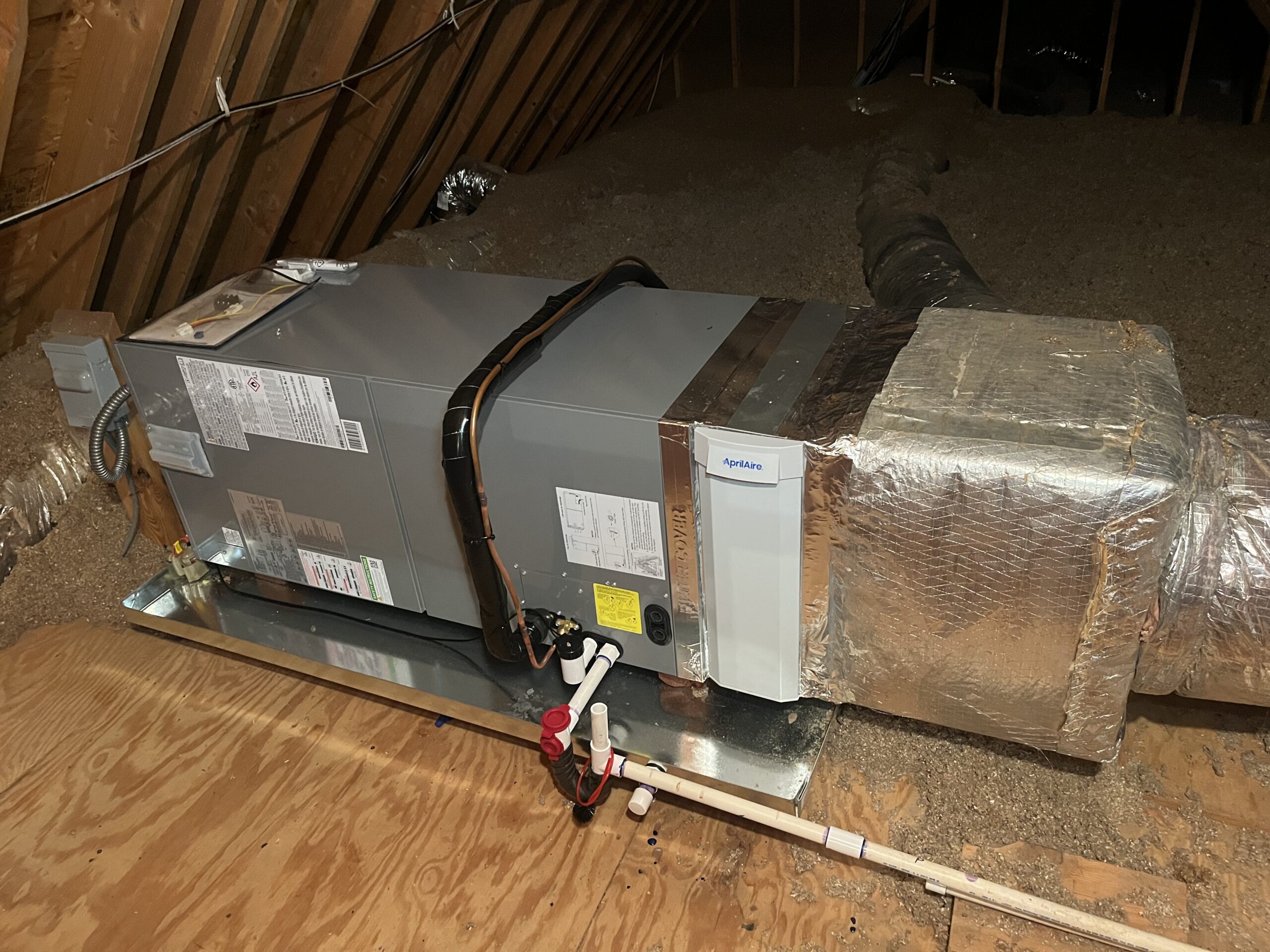 About Us: A central HVAC air handler unit installed in an attic, with insulated ductwork and pipes connected, sitting on a wooden platform.