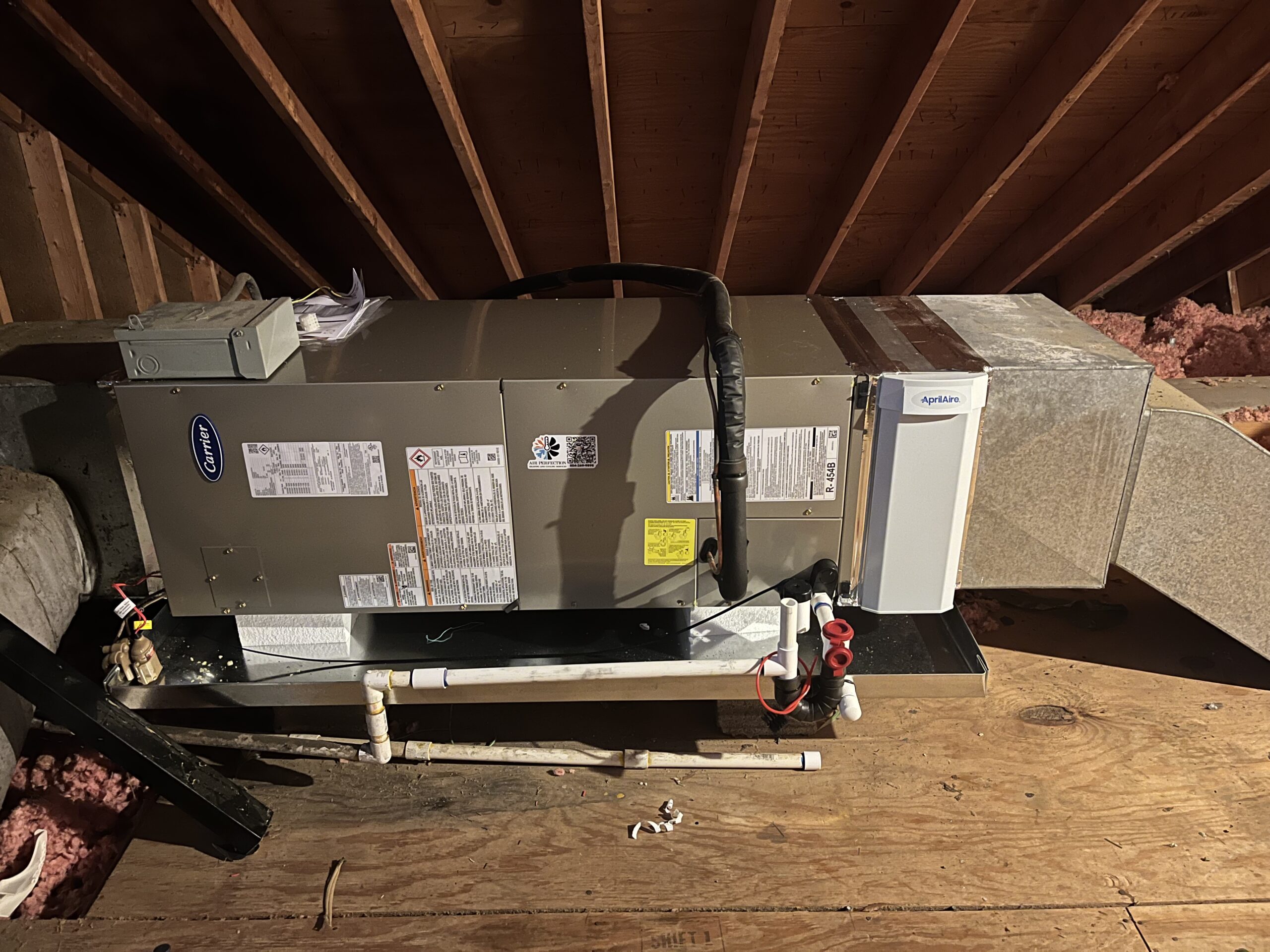 About Us: Our HVAC air handler unit, complete with connected ductwork and pipes, is expertly installed on a wooden platform in an attic featuring exposed insulation and beams.