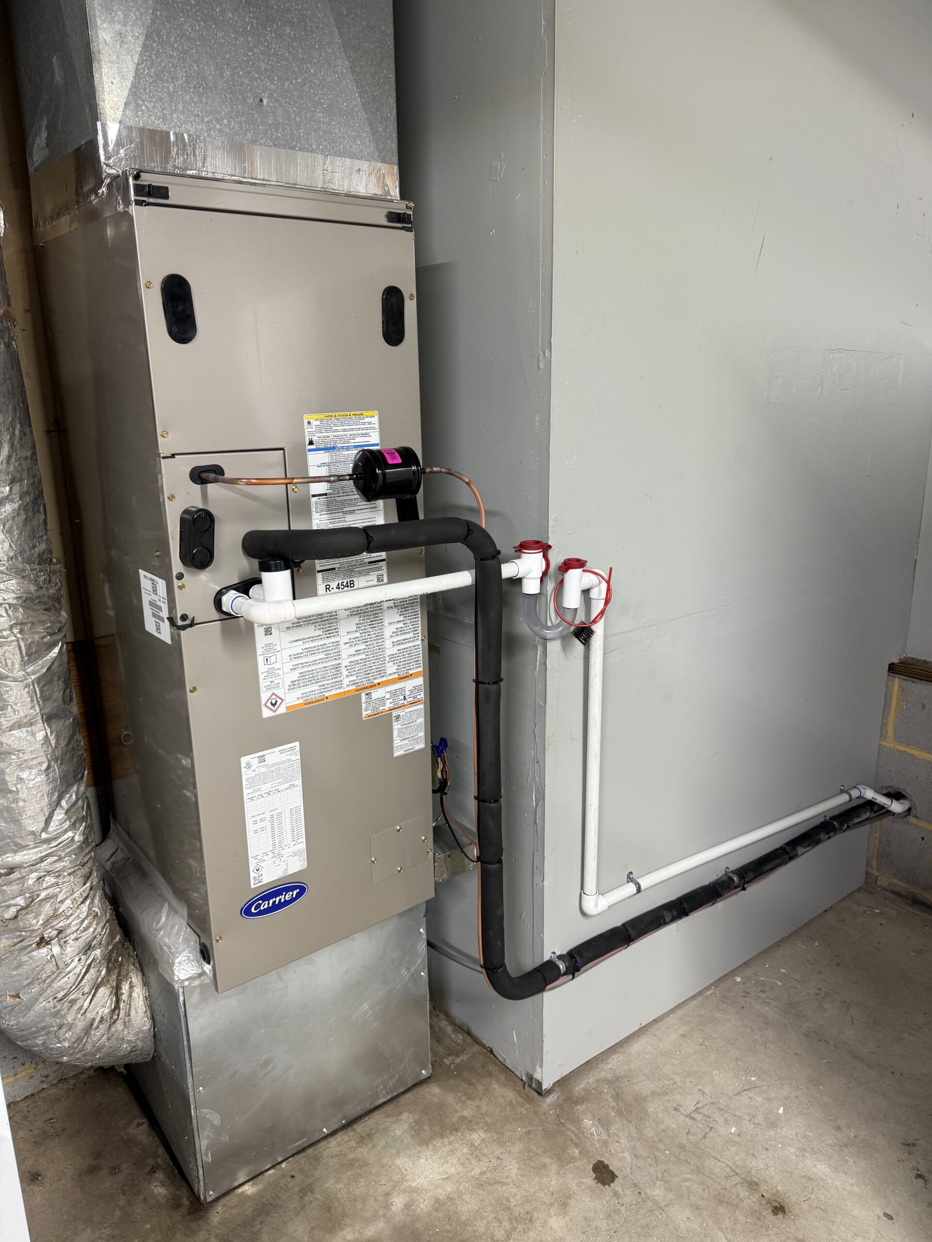 A Carrier HVAC unit with ductwork and insulated pipes is installed next to a gray utility wall in a garage or utility room—learn more about our installation expertise on the About Us page.