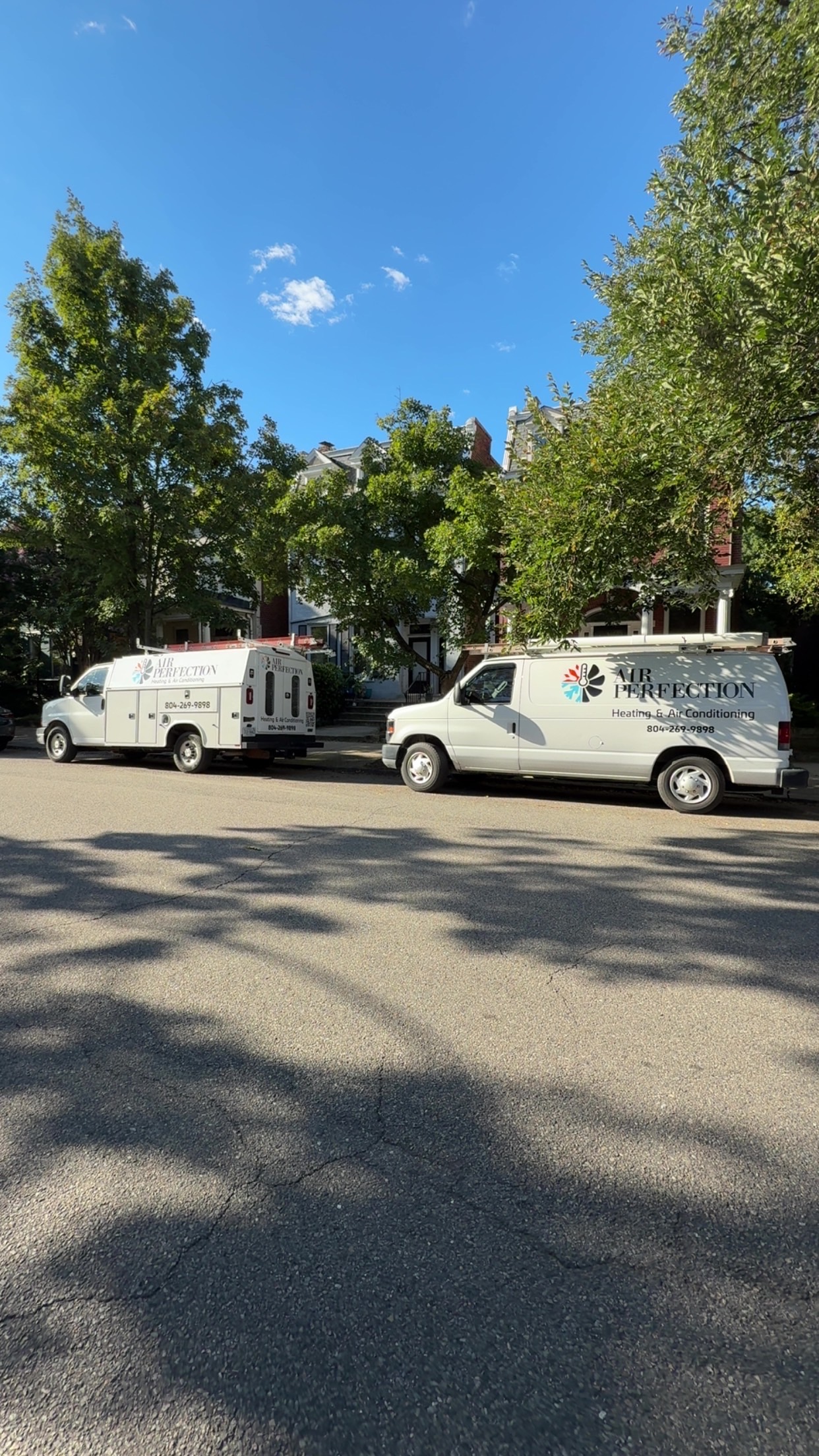 Two white utility vans with The Inspection company logos, featured in our About Us section, are parked on a residential street lined with trees and houses under a clear blue sky.