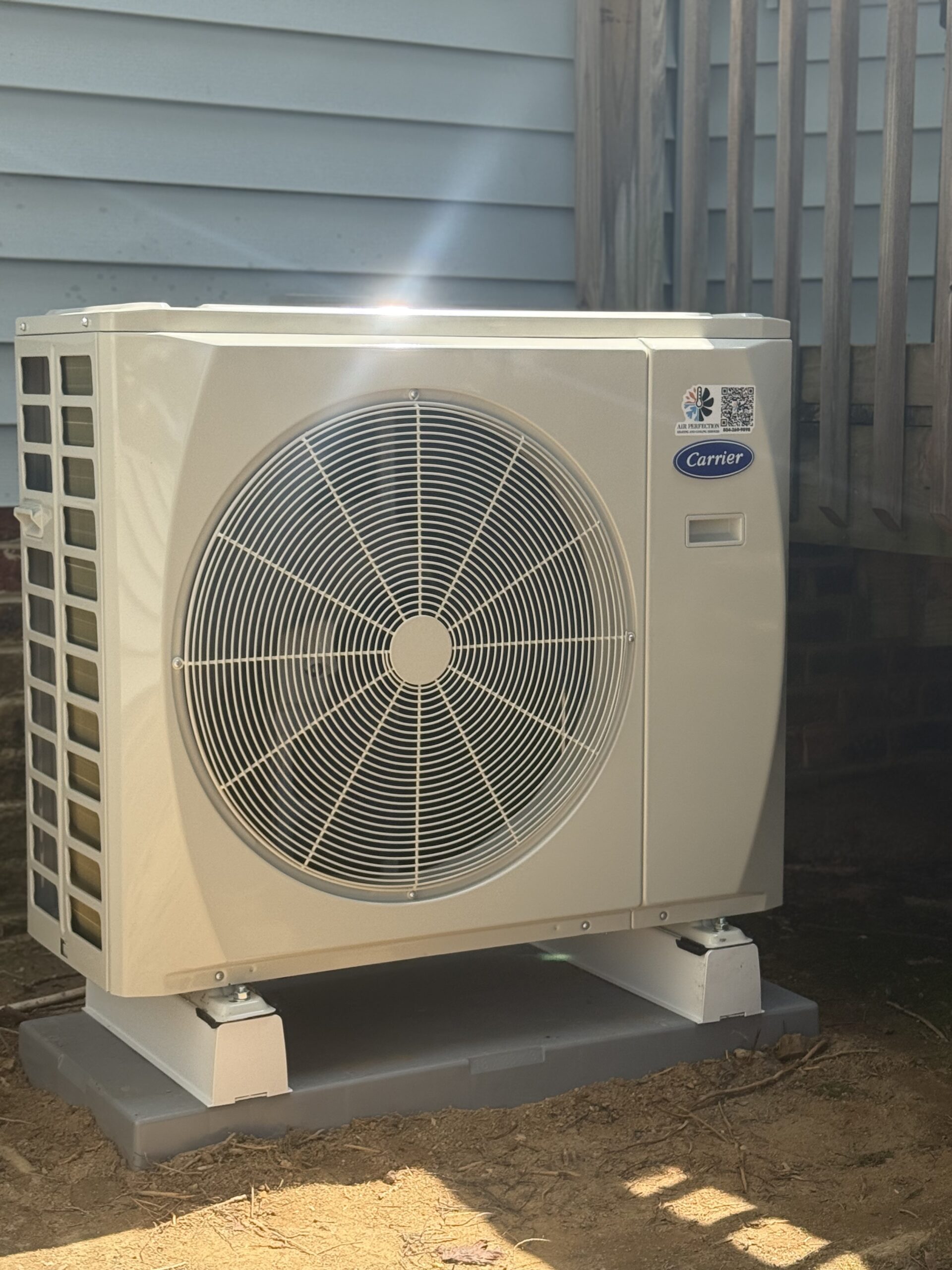 Outdoor air conditioning unit installed on a platform next to a house with siding and a wooden staircase in the background. Learn more About Us and our installation services for your home comfort needs.