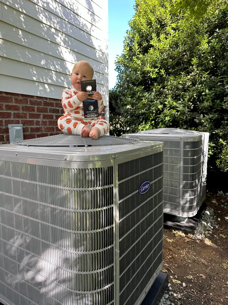 A baby in patterned pajamas sits on top of an outdoor air conditioning unit, holding a small rectangular object, with another unit and greenery nearby.