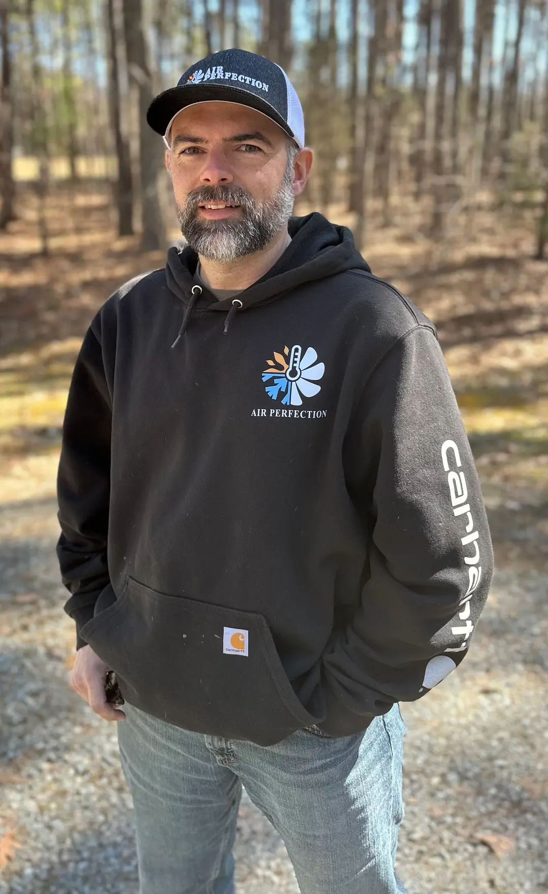 A man with a beard wearing a black "Air Perfection" hoodie and matching cap stands outdoors on a sunny day in a wooded area, embodying the casual, adventurous spirit of our About Us story.